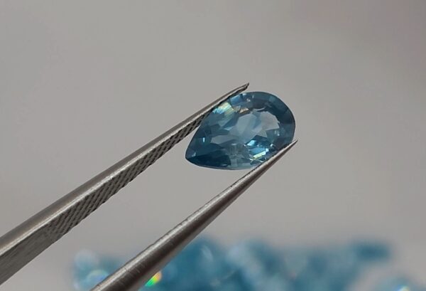 Pear-shaped faceted natural zircon gemstone held with tweezers by a gemologist, showing clarity and sparkle